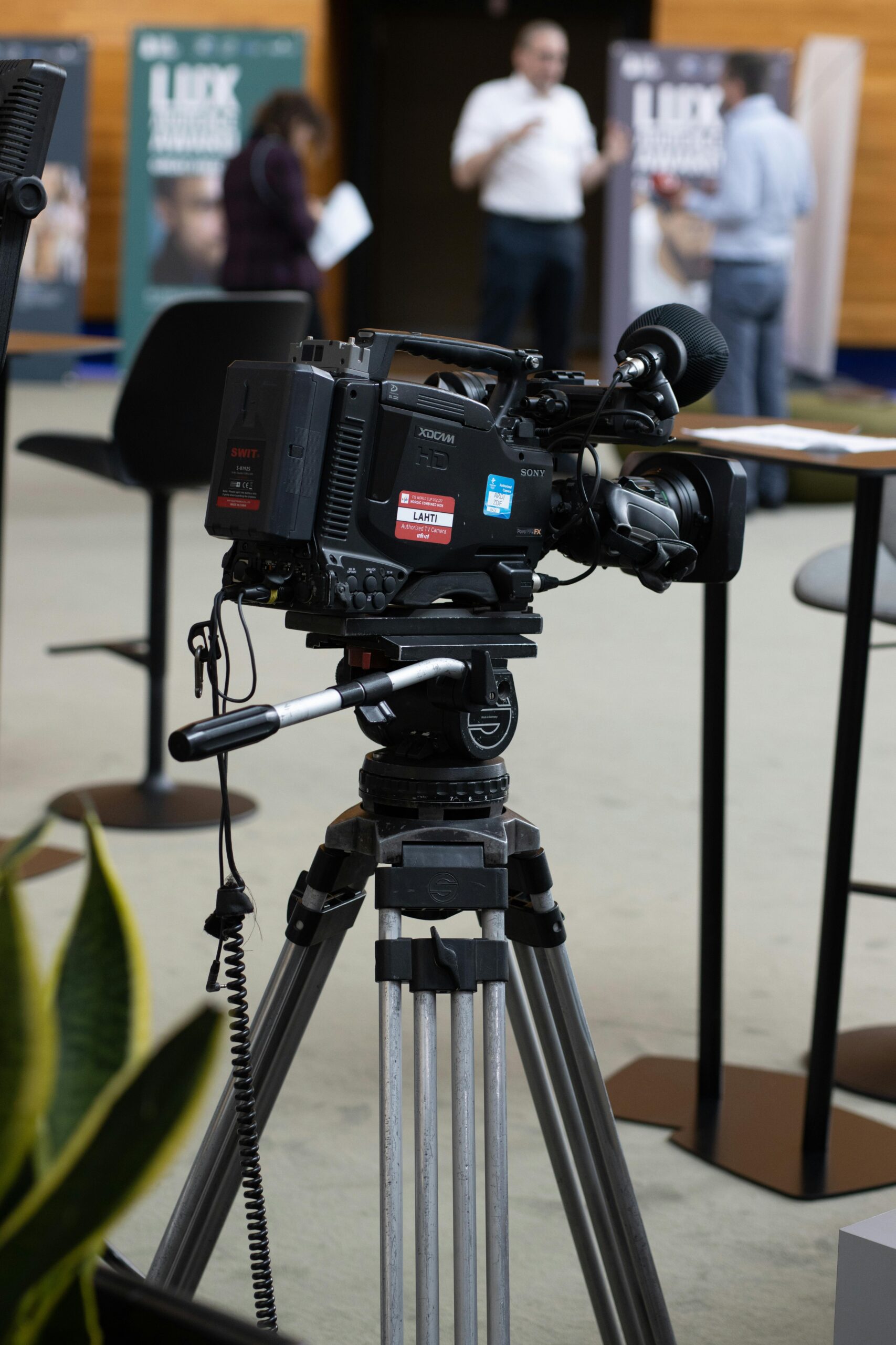 A video camera on a tripod in a modern office setting with people in the background at Strasbourg event.
